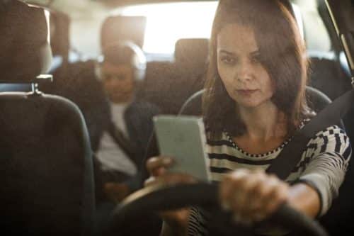 A woman using her phone while driving with a passenger in the back seat