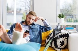 Young man with cast on left leg sitting on couch next to crutch