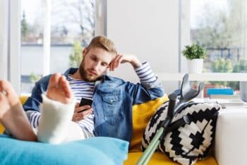 Young man with cast on left leg sitting on couch next to crutch