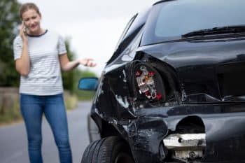 Image of a woman on the phone with a crashed black car with front end damage
