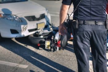 Rear view of a police officer at the scene of an accident between a white car and motorcycle.