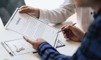 A man holding an insurance policy he’s about to sign. Another form and car keys on the desk.