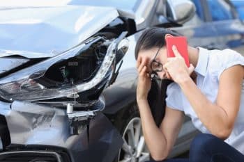 Woman on the phone holding her head next to wrecked silver car