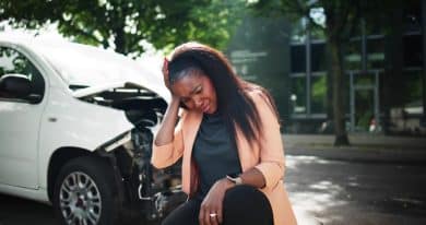 Young black woman holding her head while sitting in front of a white wrecked car