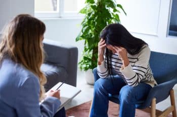 Upset woman with her head in hands sitting on chair discussing PTSD with a female therapist