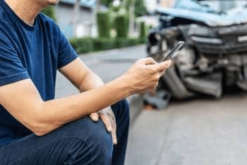 A man calling an attorney after an accident with a damaged car in the background